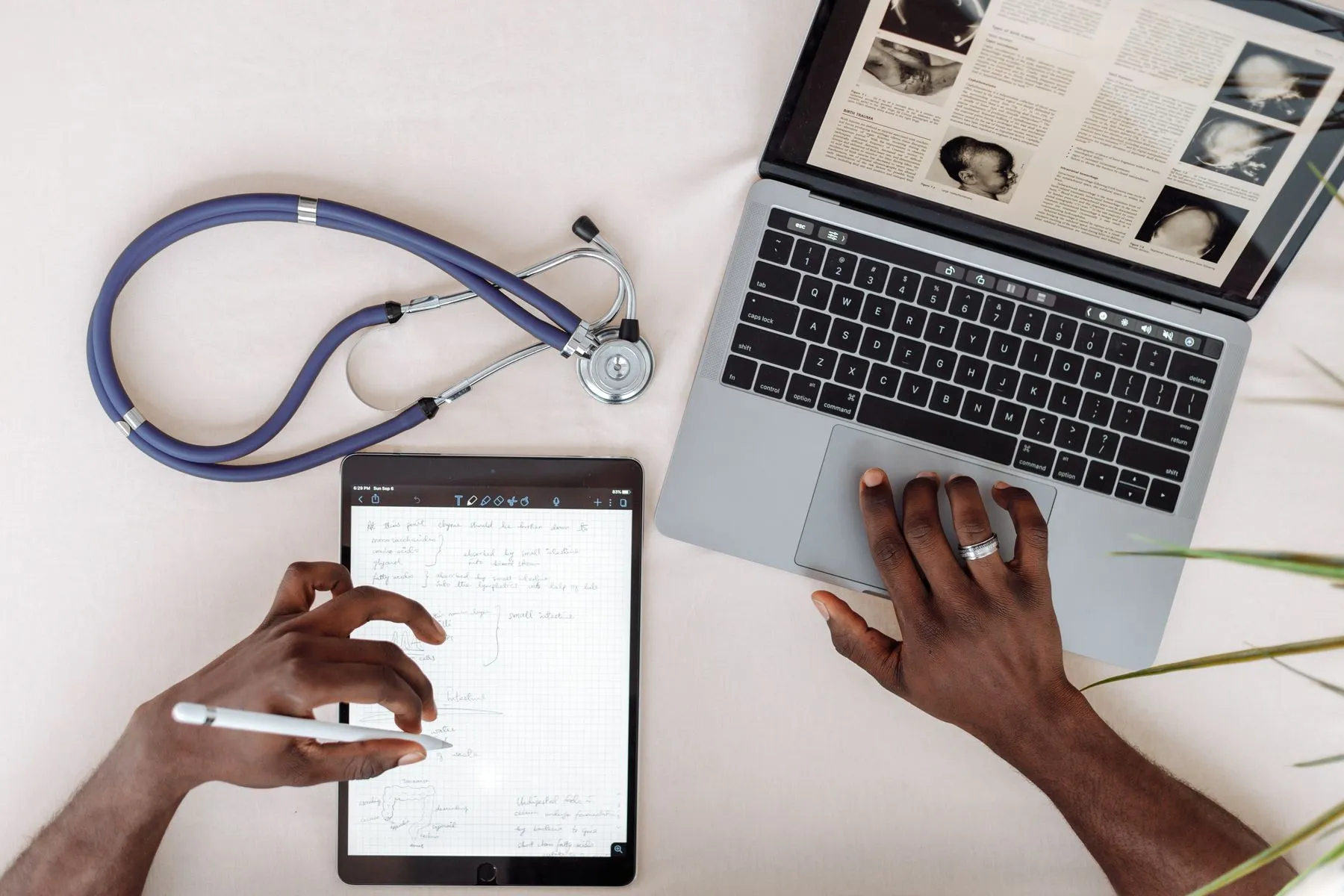Hands working on a tablet and laptop next to a stethoscope